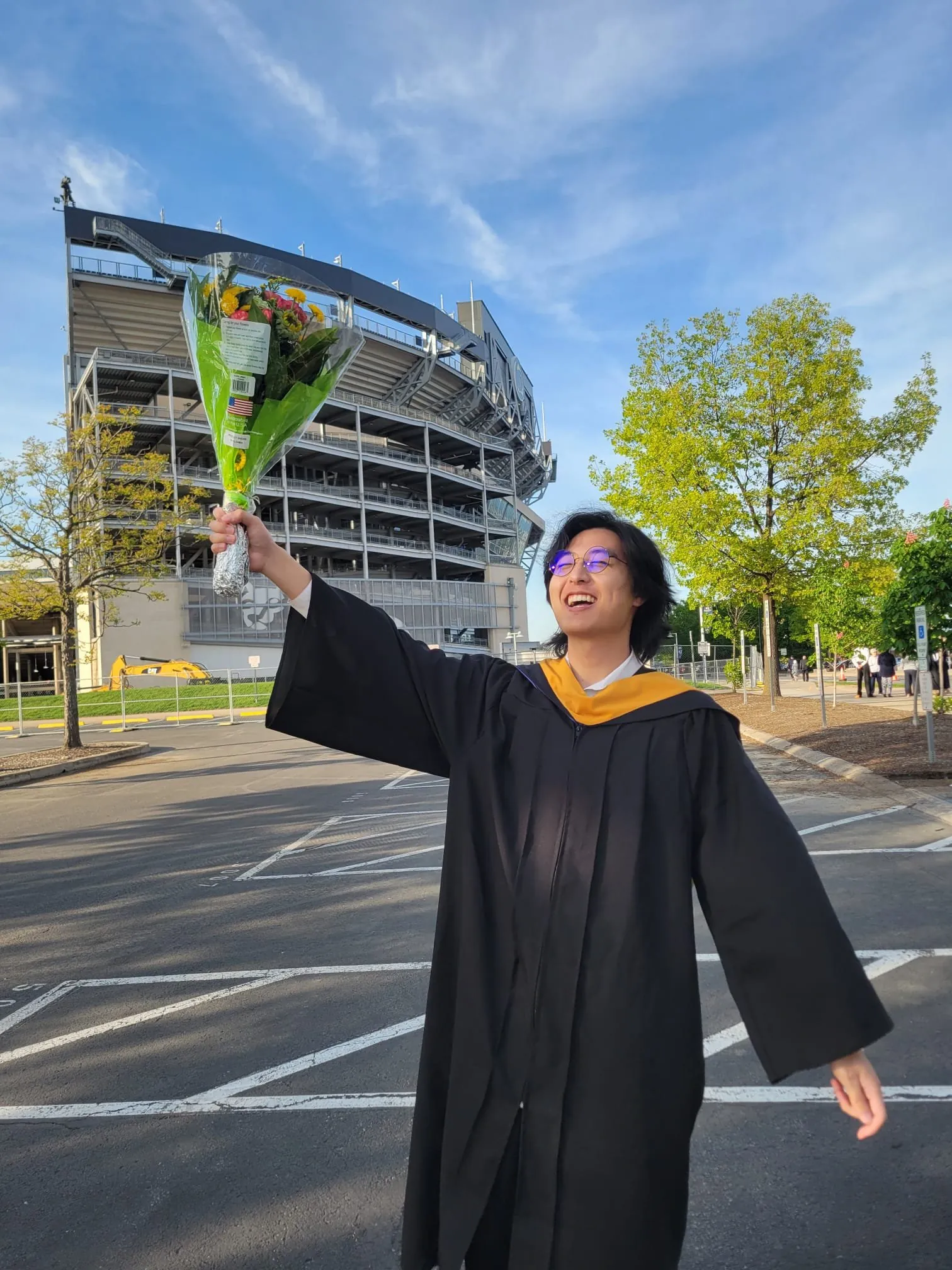 Jeffrey in a graduation gown, smiling and holding a bouquet of flowers above his head outside a stadium.